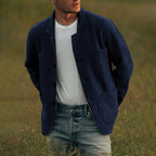 Homme portant une veste bleue en laine, t-shirt blanc et jeans, debout dans un champ verdoyant. Mode masculine décontractée, style automnal.
