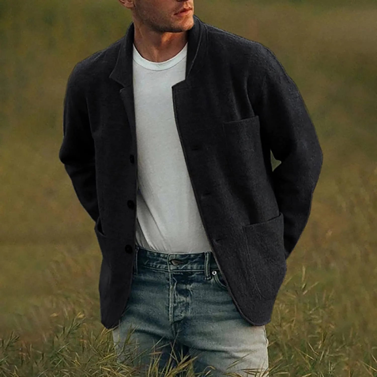 Homme portant une veste noire élégante sur un t-shirt blanc et un jean, debout dans un champ verdoyant. Mode masculine, style décontracté chic.