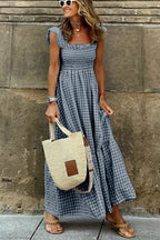 Femme en robe longue à carreaux bleus, portant un sac en paille et des sandales, souriante devant un mur en pierre. Mode estivale décontractée.
