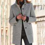 Femme portant un manteau gris élégant en laine, col relevé, sur un pull noir, dans une rue urbaine. Mode automne-hiver, style chic et moderne.