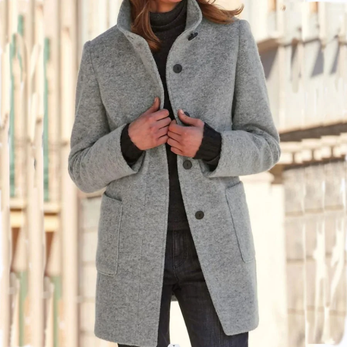 Femme portant un manteau gris élégant en laine, col relevé, sur un pull noir, dans une rue urbaine. Mode automne-hiver, style chic et moderne.