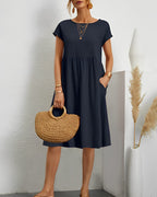 Robe midi noire élégante avec manches courtes, portée par une femme tenant un sac en paille. Décor minimaliste avec vase et plantes séchées.