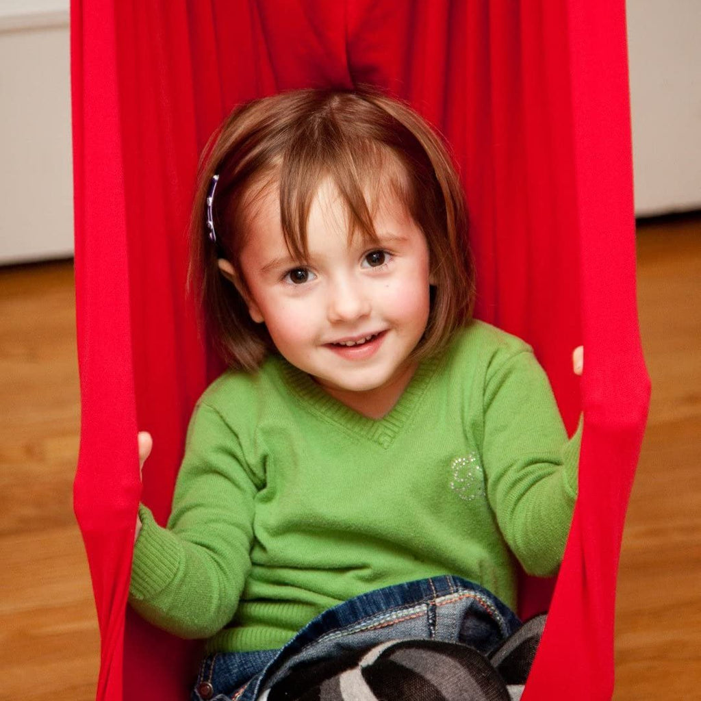 Jeune fille souriante dans un hamac rouge, portant un pull vert et un jean, sur un sol en bois. Enfant, détente, intérieur, jeu.