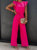 Combinaison rose vif élégante avec manches à volants, ceinture nouée, pochette beige et sandales à talons, tenue chic pour soirée ou événement.