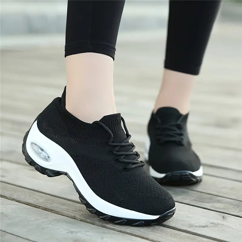 Chaussures de sport noires pour femme avec semelle épaisse blanche, portées sur un sol en bois. Confortables et tendance, idéales pour la marche.