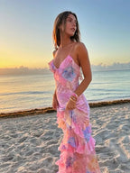 Femme en robe longue colorée sur plage au coucher du soleil, regardant vers l'horizon. Mer calme et ciel orangé en arrière-plan.