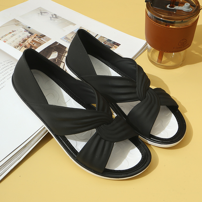 Sandales noires élégantes avec nœud croisé sur fond jaune, posées sur un magazine ouvert, à côté d'un gobelet en plastique transparent.