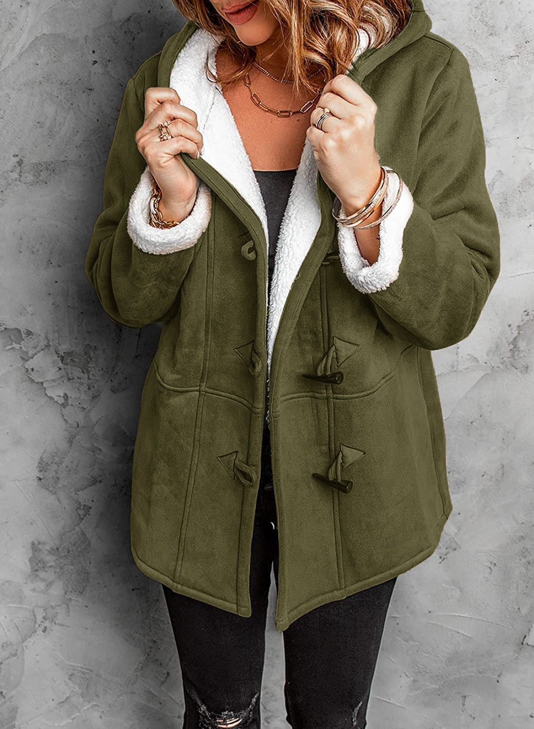 Femme portant un manteau vert à capuche doublé de fourrure blanche, style décontracté, sur fond de mur gris. Mode hiver, tendance féminine.