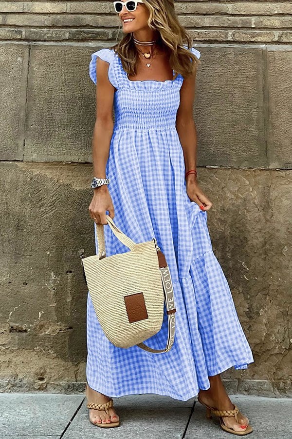 Femme en robe vichy bleue, lunettes de soleil blanches, sac en paille, sandales, posant devant un mur en pierre. Mode estivale chic et décontractée.