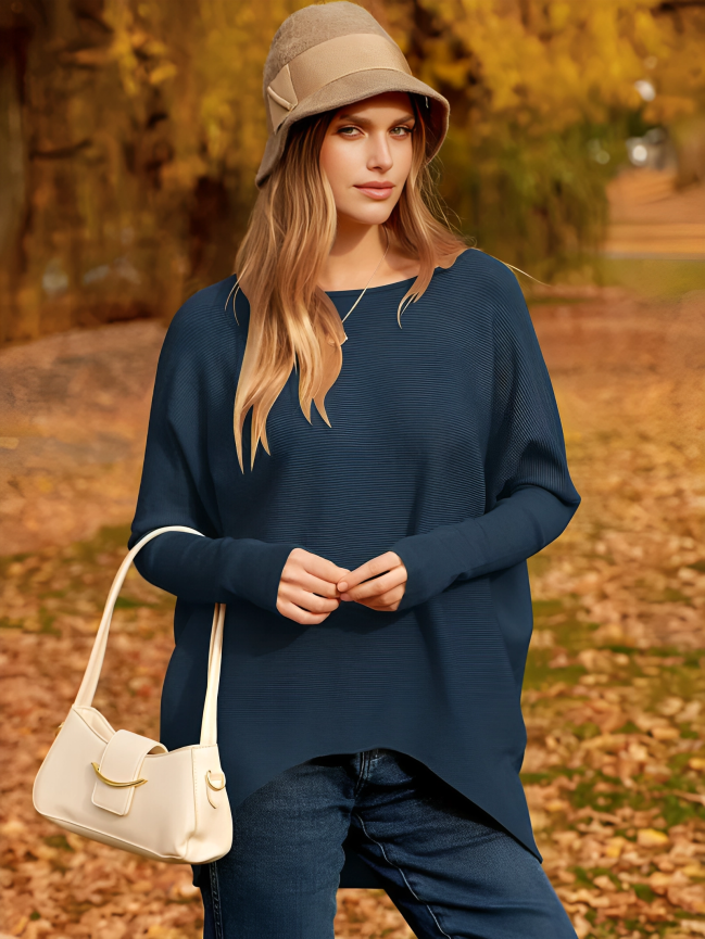 Femme en pull bleu et chapeau beige, posant dans un parc automnal avec feuilles jaunes. Mode automne, style décontracté, sac à main blanc.