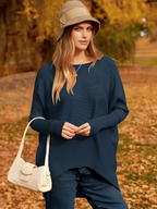 Femme en pull bleu et chapeau beige, posant dans un parc automnal avec feuilles jaunes. Mode automne, style décontracté, sac à main blanc.