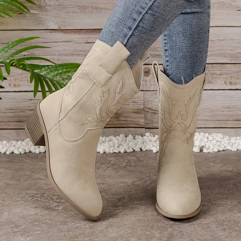 Bottes western beiges en cuir avec broderie florale, portées avec un jean ajusté, posées sur un sol en pierre, décor végétal en arrière-plan.