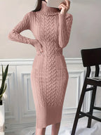 Robe pull rose en tricot torsadé, manches longues, col roulé, ajustée, élégante, posée près d'une chaise noire et d'une plante verte.