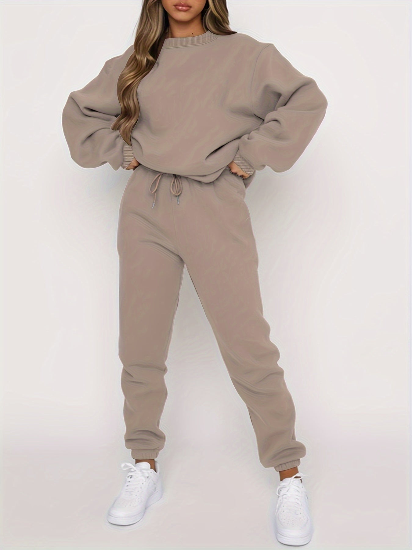 Femme portant un ensemble de survêtement beige, composé d'un sweat ample et d'un pantalon de jogging, avec des baskets blanches. Mode décontractée.