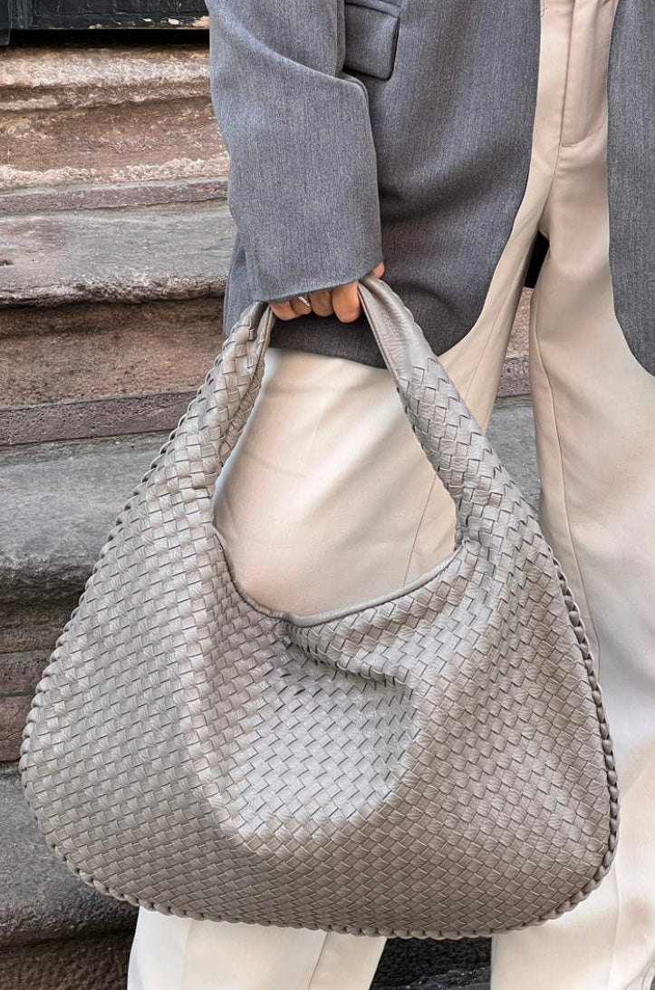 Sac à main tissé gris argenté, porté par une personne en pantalon blanc et veste grise, sur des marches en pierre. Mode élégante et tendance.