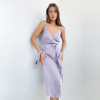 Femme portant une robe lilas en lin, style portefeuille, bretelles fines, posant contre un mur blanc. Mode féminine élégante et estivale.