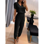 Femme portant une combinaison noire élégante, posant dans un salon moderne avec des meubles noirs et une plante verte. Mode féminine chic et tendance.