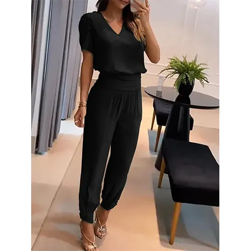 Femme portant une combinaison noire élégante, posant dans un salon moderne avec des meubles noirs et une plante verte. Mode féminine chic et tendance.