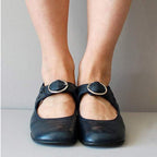 Chaussures Mary Jane noires avec boucle argentée portées par une personne debout sur un sol gris. Mode féminine, chaussures classiques élégantes.