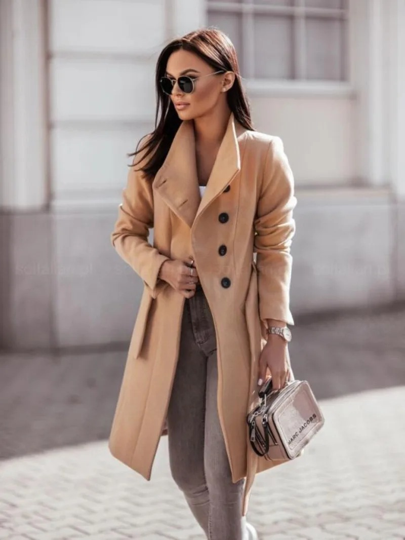 Femme élégante portant un manteau camel, lunettes de soleil noires et sac à main argenté, marchant en extérieur. Mode automne chic et tendance.