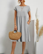 Robe grise élégante portée par une femme, accessoirisée d'un sac en osier et de bijoux dorés, posant dans un intérieur minimaliste avec plantes décoratives.
