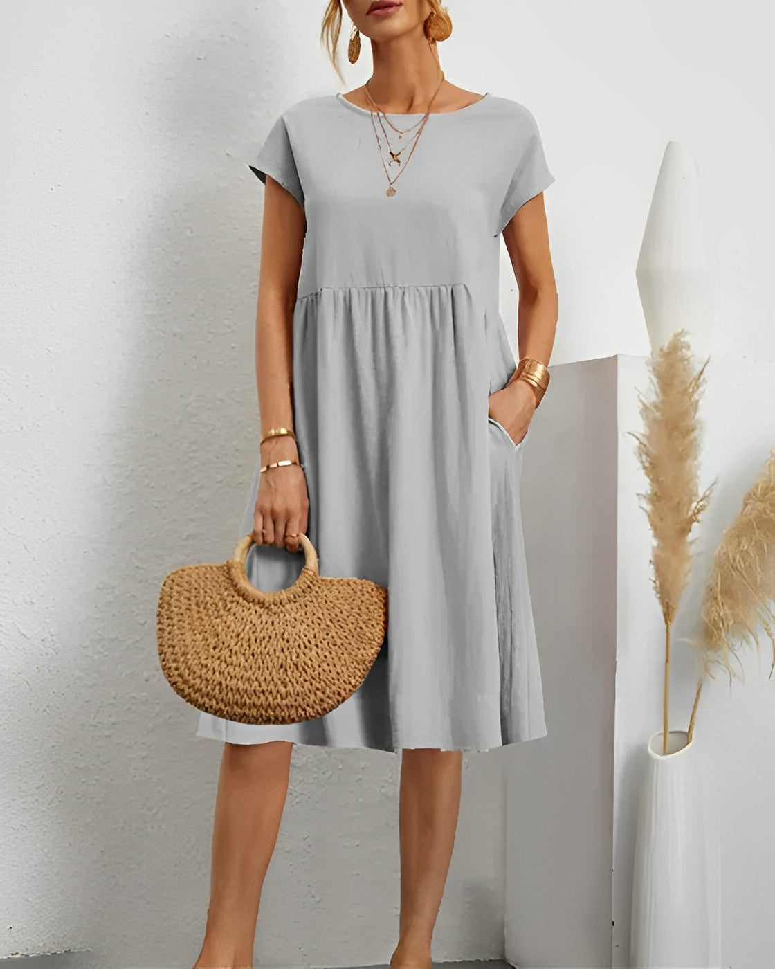 Robe grise élégante portée par une femme, accessoirisée d'un sac en osier et de bijoux dorés, posant dans un intérieur minimaliste avec plantes décoratives.