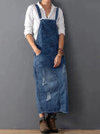 Femme portant une robe salopette en jean déchiré, chemise blanche, bottines marron, posant contre un mur gris. Mode décontractée, tendance denim.