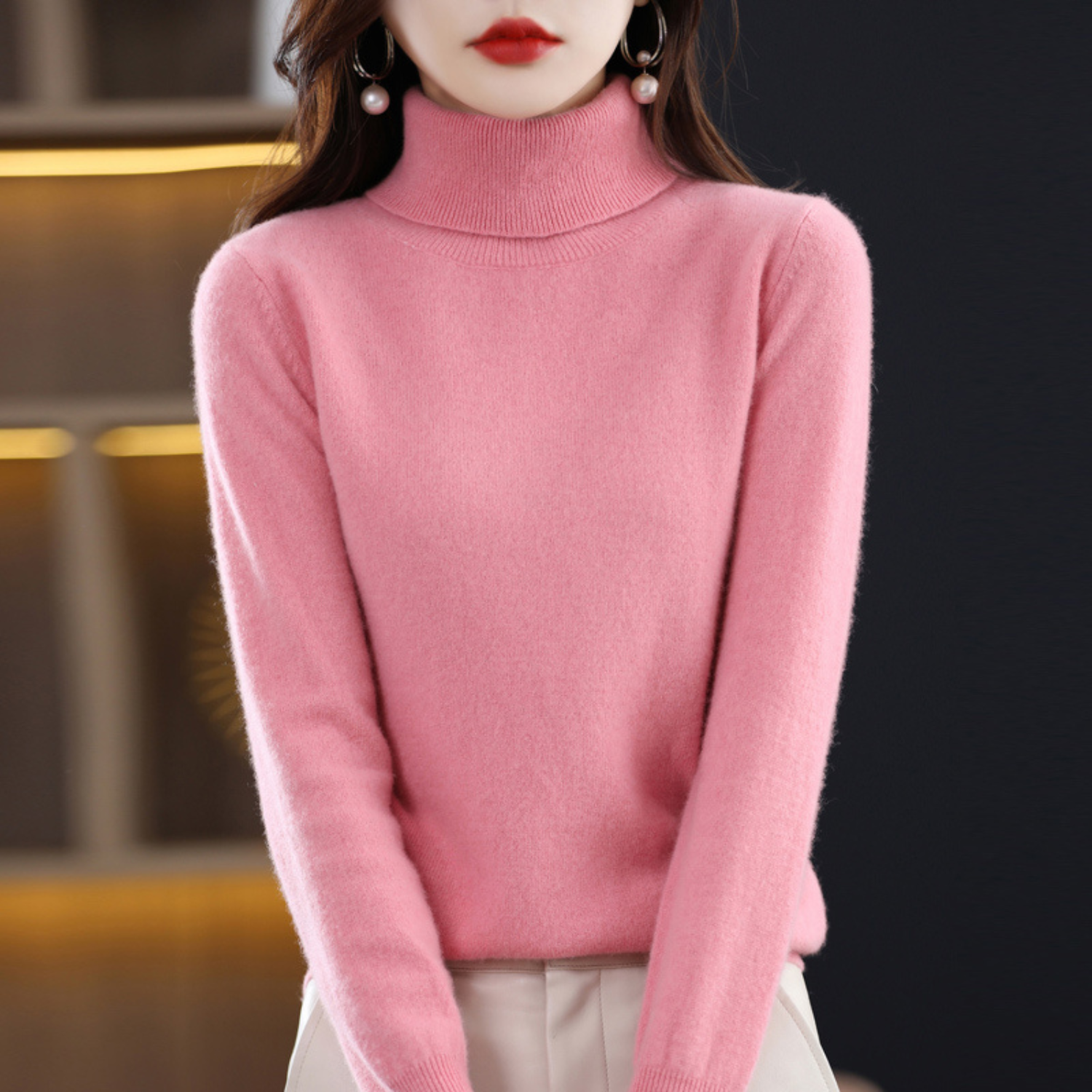 Femme portant un pull rose en cachemire à col roulé, élégante et moderne, idéal pour la mode automne-hiver. Accessoires: boucles d'oreilles perles.