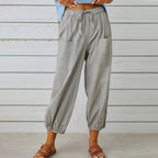 Pantalon ample gris avec poches avant, taille élastique et boutons décoratifs aux chevilles, porté par une femme sur fond de planches en bois.