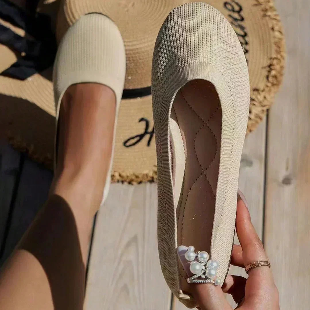Ballerine beige en tissu texturé, tenue à la main, avec perles décoratives, posée sur un chapeau de paille sur une terrasse en bois.