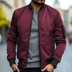 Homme portant un blouson bomber bordeaux, t-shirt gris, dans une rue floue. Mode masculine, veste tendance, style urbain décontracté.