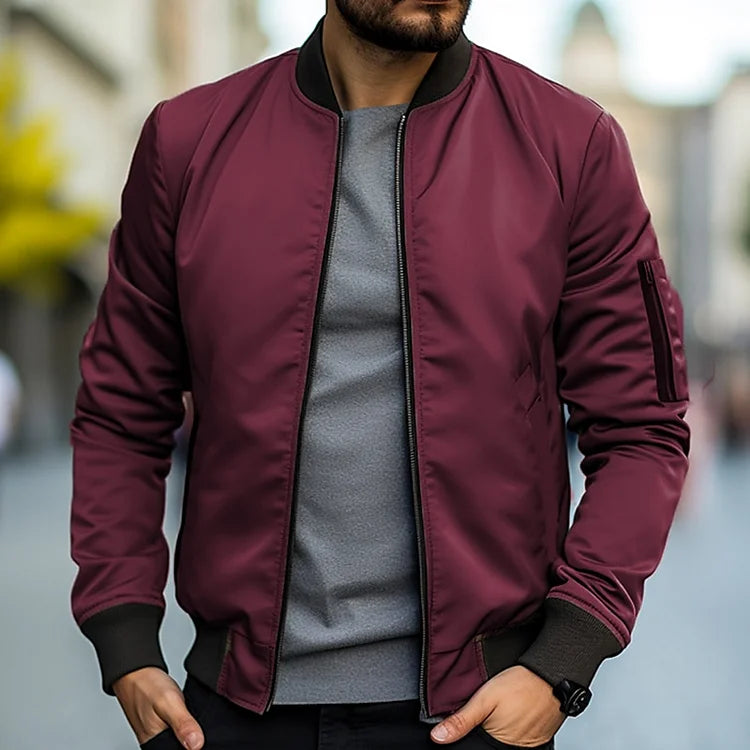 Homme portant un blouson bomber bordeaux, t-shirt gris, dans une rue floue. Mode masculine, veste tendance, style urbain décontracté.