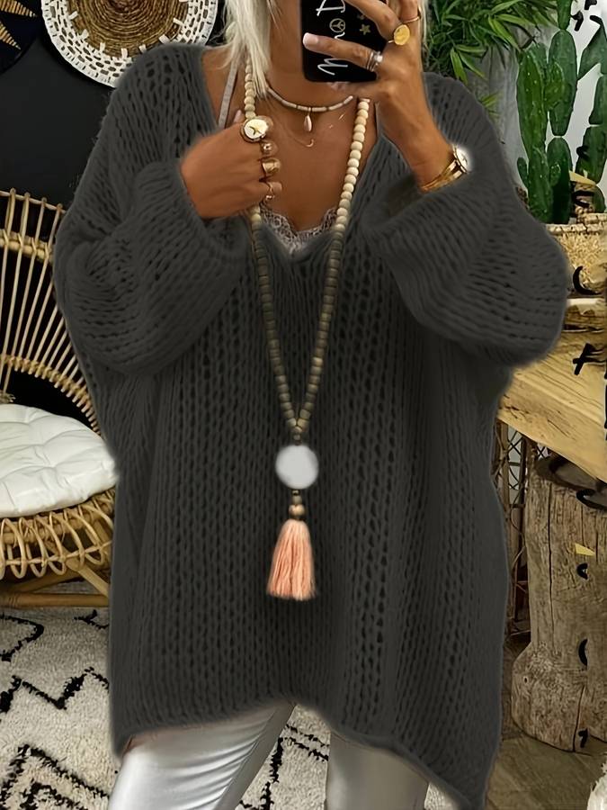 Femme portant un pull oversize noir, collier long avec pompon, leggings blancs. Décor intérieur avec chaise en rotin et plantes vertes.