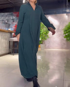 Femme portant une robe longue verte, marchant dans un intérieur moderne avec plantes et murs texturés. Mode élégante, tenue décontractée chic.