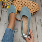 Ballerines vertes en tricot avec perles, tenues par une main, posées sur un sol en bois avec un sac en osier et des fleurs jaunes en arrière-plan.