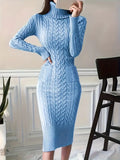Robe pull bleu en tricot torsadé, manches longues, col roulé, portée par une femme debout dans un intérieur moderne avec chaise et plante.