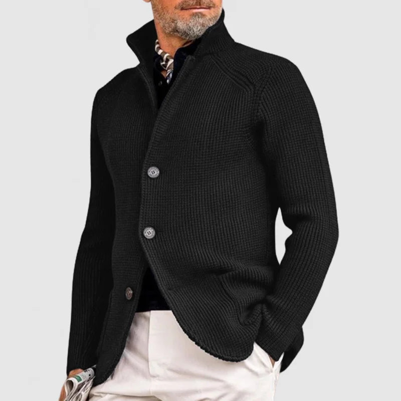 Homme portant un cardigan noir élégant avec boutons, col relevé, sur chemise noire et pantalon beige. Mode masculine chic et moderne.