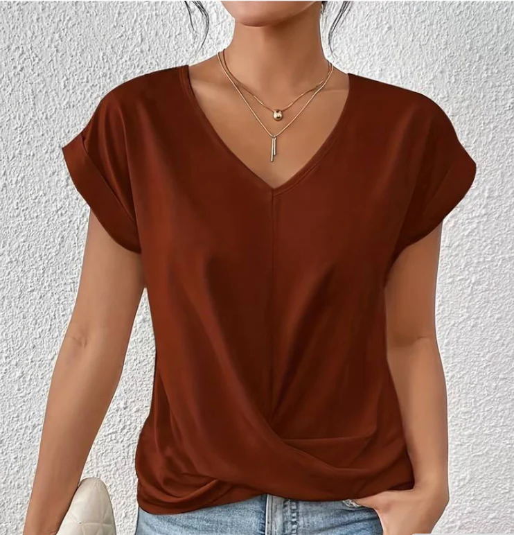 Femme portant un t-shirt marron à col en V, manches courtes, avec colliers dorés, posant devant un mur blanc texturé. Mode décontractée élégante.