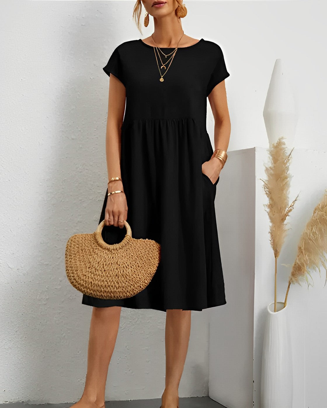 Robe noire élégante femme, manches courtes, col rond, avec poches. Accessoires: collier doré, sac en paille. Décor minimaliste, vase pampas.