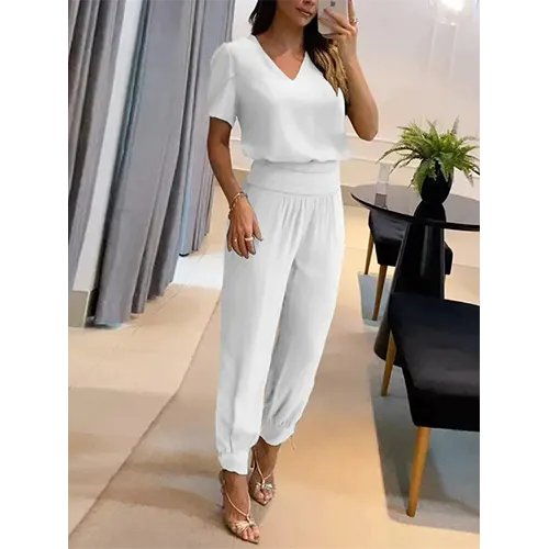 Femme portant une combinaison blanche élégante, debout dans un salon moderne avec des rideaux gris et une plante verte sur une table noire.