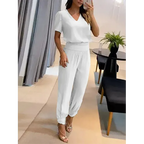 Femme portant une combinaison blanche élégante, debout dans un salon moderne avec des rideaux gris et une plante verte sur une table noire.