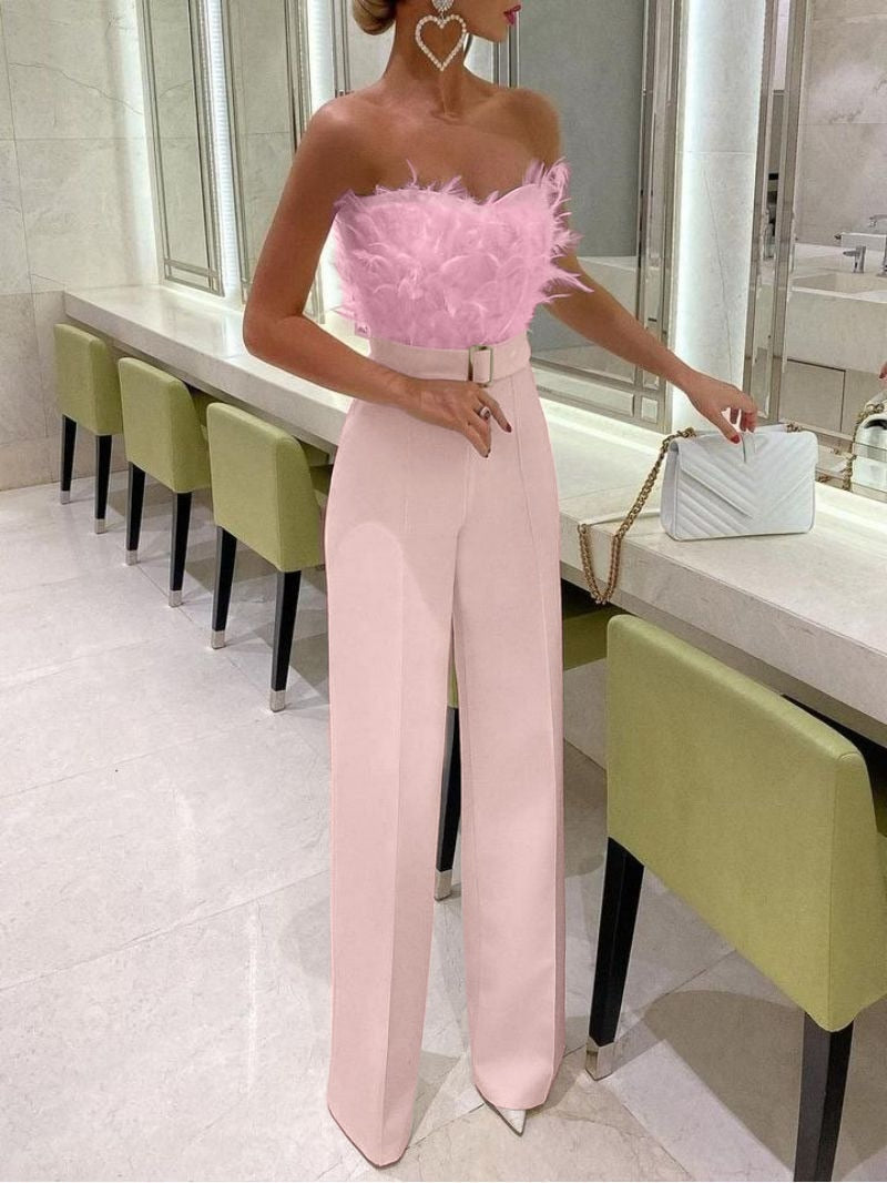 Femme en tenue élégante avec haut à plumes roses et pantalon taille haute rose, posant dans un intérieur moderne avec chaises vertes et sac blanc.
