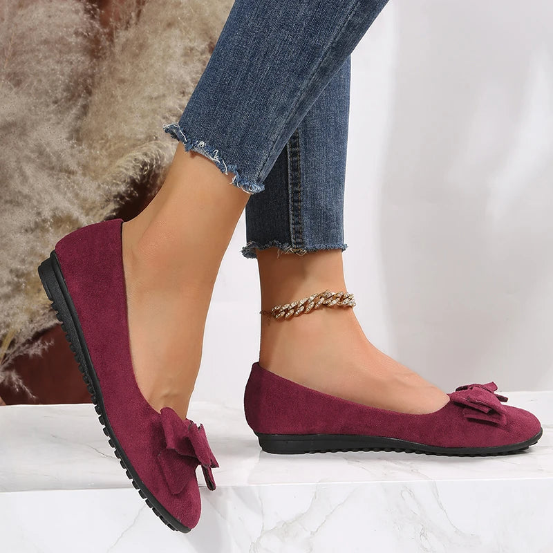 Ballerines bordeaux en daim avec nœud, portées avec un jean bleu déchiré à l'ourlet, posées sur un fond neutre. Chaussures élégantes et confortables.