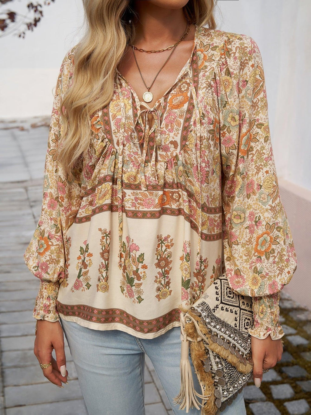 Femme portant un chemisier bohème à motifs floraux avec manches longues, collier doré, sac à main à franges, et jeans. Mode bohème chic tendance.