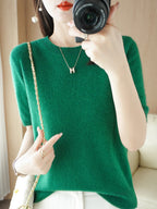 Femme en pull vert, tenant un appareil photo, portant un collier avec pendentif "H" et un sac à bandoulière doré. Mode élégante et moderne.