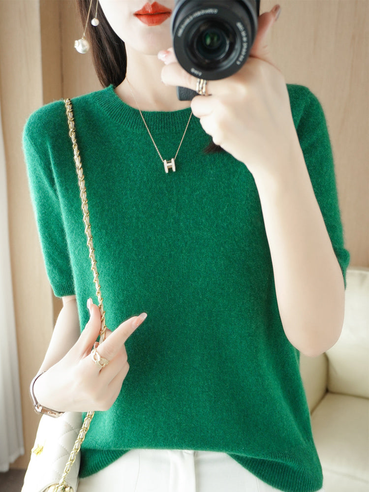 Femme en pull vert, tenant un appareil photo, portant un collier avec pendentif "H" et un sac à bandoulière doré. Mode élégante et moderne.