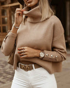 Femme portant un pull beige à col roulé avec boutons, montre élégante, sac à main marron, ceinture blanche, et pantalon blanc. Mode automne chic.
