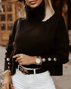 Femme en pull noir à col roulé avec boutons, montre élégante, pantalon blanc, ceinture noire, posant en extérieur urbain. Mode automne chic.