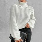 Femme portant un pull blanc en maille à col roulé, tenant un sac à main noir avec chaîne dorée, sur fond gris. Mode automne-hiver élégante.