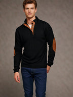 Homme portant un pull noir avec coudières marron et col boutonné, debout en jeans. Mode masculine élégante, tenue automne-hiver tendance.